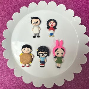 May include: Six small, colorful figurines of characters from the animated television show Bob's Burgers. The characters include Bob Belcher, Linda Belcher, Tina Belcher, Gene Belcher, Louise Belcher, and Teddy.