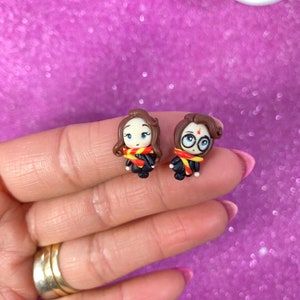 May include: A pair of handmade polymer clay earrings featuring a cartoon depiction of Harry Potter and Hermione Granger. The earrings are painted in shades of brown, yellow, red, and black.