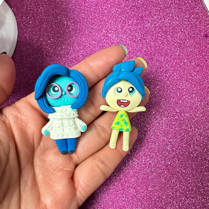 May include: Two miniature figurines of characters from the animated film "Inside Out." One figurine is blue with glasses and a white sweater. The other is yellow and green with a smiling expression and outstretched arms. The figurines are held in a hand.