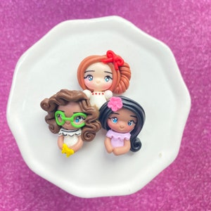 May include: Three polymer clay dolls with different hairstyles and outfits. The doll on the left has brown hair, green glasses, and a white dress. The doll in the middle has red hair, a red bow, and a white dress. The doll on the right has brown hair, a pink flower, and a purple dress.
