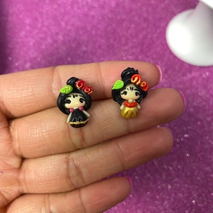 May include: Two small, colorful, handmade clay figurines of women with black hair, red and gold accents, and green leaves. The figurines are wearing traditional Mexican clothing.