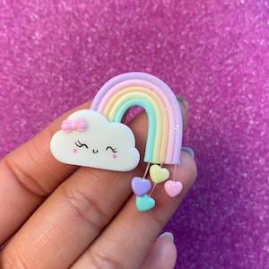 May include: A pastel rainbow with a white cloud with a pink bow and a happy face.  The rainbow has glitter and there are five pastel hearts hanging from the bottom of the rainbow.