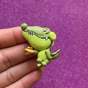 May include: A handmade, green alligator figurine with a gold-colored hand and a brown tail. The alligator has black details and a friendly expression. The figurine is made of clay and is set against a purple background.