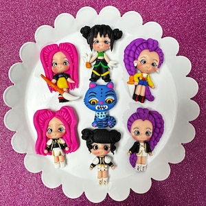 May include: A collection of handmade polymer clay figurines, including dolls with pink and purple hair, and a blue cat. The dolls wear various outfits with black, white, yellow, and green accents. The figurines are arranged on a white scalloped plate.