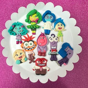 May include: A collection of colorful clay figurines of characters from the animated movie "Inside Out" arranged on a white scalloped-edge plate. The figurines feature various emotions, including Joy, Sadness, Anger, Fear, and Disgust, with detailed facial expressions and clothing.