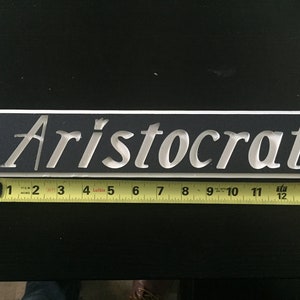 May include: A white rectangular sign with the word "Aristocrat" written in a silver, cursive font on a black background.