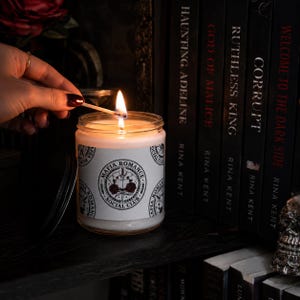 Pode incluir: Uma vela em pote de vidro transparente com um rótulo branco apresentando um logotipo circular com o texto Mafia Romance Social Club, centrado em torno de uma ilustração de um livro, espada e rosas. A vela está acesa com uma chama.