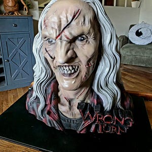 May include: A detailed bust of a horror character with long, wavy white hair, pale skin, and visible facial scars. The figure wears a red and black plaid shirt and features the text WRONG TURN in bold, dark lettering on the lower chest area.