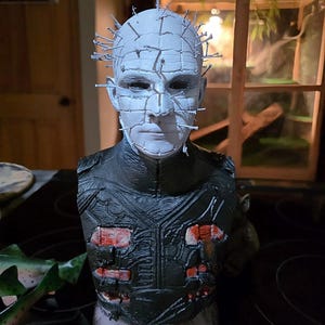 May include: A bust of a character with a pale face, a grid-like pattern, and metal pins protruding from the head. The figure wears a black, textured garment with red accents, evoking a horror theme.