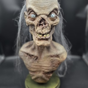 May include: A detailed bust of a gaunt, humanoid creature with textured, weathered skin, sunken features, and pale blue eyes. The figure has long, thin, wispy gray hair and a skeletal chest, mounted on a circular, muted green base.