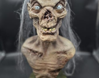 May include: A detailed bust of a gaunt, humanoid creature with textured, weathered skin, sunken features, and pale blue eyes. The figure has long, thin, wispy gray hair and a skeletal chest, mounted on a circular, muted green base.