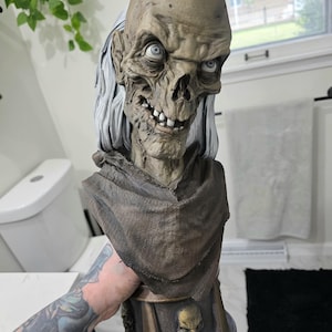 May include: A detailed bust of a skeletal figure with sunken eyes, a wide grin showing teeth, and long white hair. The figure wears a textured brown cowl over its shoulders and sits atop a pedestal base featuring a small skull emblem in the center.