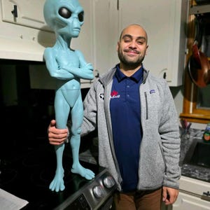 May include: A light blue alien figurine with large black eyes and a slender build. The figurine is standing on a black surface. The alien is being held by a person wearing a gray jacket and a blue shirt.