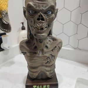 May include: A brown, weathered bust of the Crypt Keeper character with sunken eyes, visible teeth, and textured, decaying skin. The figure is mounted on a rectangular base featuring the green, raised text "TALES FROM THE CRYPT" on the front.