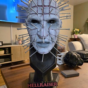 May include: A detailed bust of the character Pinhead from the Hellraiser film series. The sculpture features a pale face with nails protruding from the head. The base is black, with the word "HELLRAISER" in red.