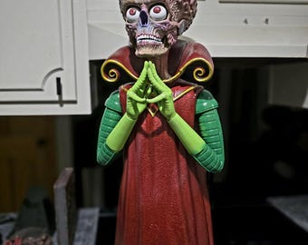 May include: A Martian figure from Mars Attacks with a large exposed brain, bulging red eyes, and a skeletal face. The figure wears a long red robe with a high gold-trimmed collar and green sleeves, with hands pressed together in a prayer-like gesture.