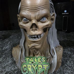 May include: A tan-colored bust of the Crypt Keeper character with sunken eyes, prominent teeth, and long gray hair. A green and black sign with the text TALES FROM THE CRYPT is attached to the front of the chest area of the sculpted figure.