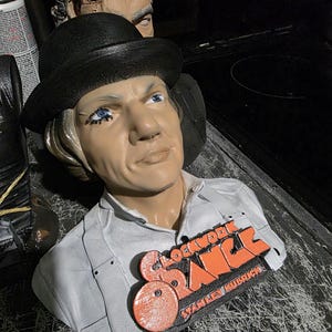 May include: A bust of a character from the film "A Clockwork Orange" wearing a black bowler hat and white shirt. The bust has a "Clockwork Orange" logo in orange and black. Another bust is visible in the background.