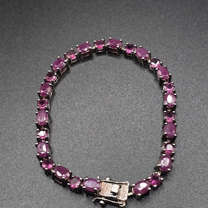 May include: A tennis bracelet featuring a series of oval-cut, deep pinkish-red gemstones set in a metallic frame. The bracelet is secured with a rectangular box clasp mechanism that includes a safety latch, creating a continuous circular loop.
