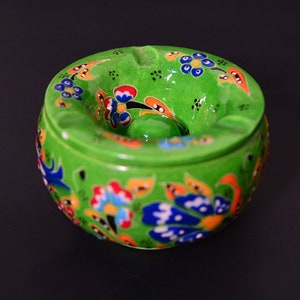 Könnte beinhalten: Ein runder, hellgrüner Keramik-Aschenbecher mit handgemaltem Blumenmuster in Blau, Rot, Orange und Schwarz. Der Aschenbecher verfügt über eine mittige Erhöhung und mehrere Einkerbungen am Rand zum sicheren Ablegen von Gegenständen.