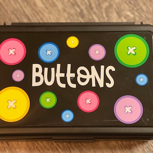 May include: A black plastic button box with a colorful button design. The box has the word "Buttons" printed on the front in white.