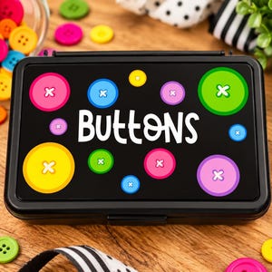 May include: A rectangular black storage box with the word Buttons printed in white on the lid, surrounded by colorful circular button graphics in pink, blue, yellow, and green, with a glass jar of loose buttons nearby.