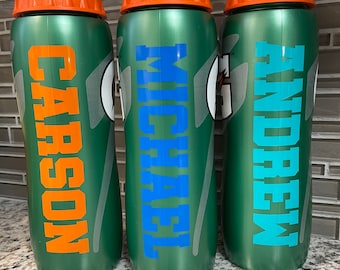 May include: Three green plastic sports water bottles with orange screw-top lids. Each bottle features a vertical Gatorade logo and a personalized name in block lettering: Carson in orange, Michael in blue, and Andrew in light blue.
