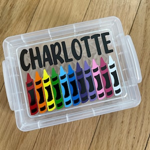 May include: A clear plastic storage container with a black label that says "CHARLOTTE" and a row of colorful crayons inside.