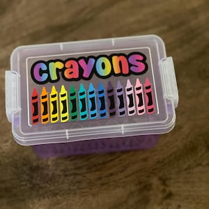 May include: A clear plastic box with a purple base and a clear lid. The lid features the word "crayons" in rainbow colors above a row of colorful crayon illustrations. The box has a small latch on each side.