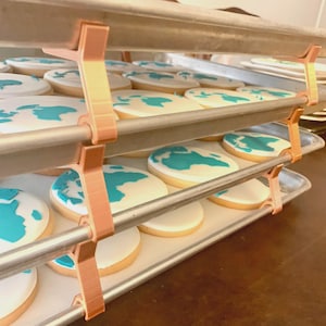 May include: A rack of iced cookies, each decorated with a blue map of the world on a white base. The cookies sit on metal trays, supported by copper-coloured brackets. The cookies are round and appear to be freshly baked.