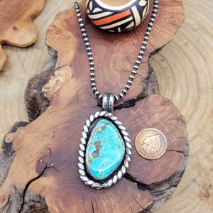 May include: A silver chain necklace with a turquoise pendant. The pendant is set in a silver frame with a rope design. The necklace is displayed on a wooden surface with a small coin.