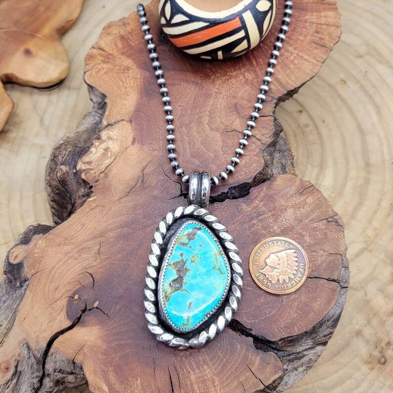 May include: A silver chain necklace with a turquoise pendant. The pendant is set in a silver frame with a rope design. The necklace is displayed on a wooden surface with a small coin.