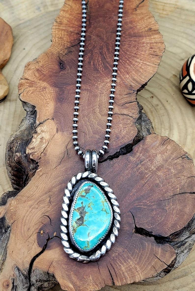 May include: A silver chain necklace with a turquoise stone pendant. The pendant is oval shaped and has a silver rope design around the edge.