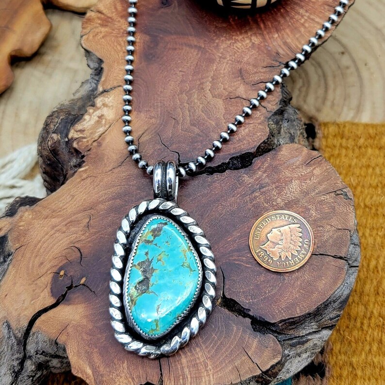 May include: A silver chain necklace with a turquoise stone pendant. The pendant is set in a silver frame with a rope design. The necklace is on a wooden surface.