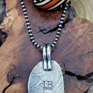 May include: A silver pendant necklace with a textured oval pendant. The pendant has a silver bail and the initials "UB" stamped on the back. The necklace is on a silver chain.
