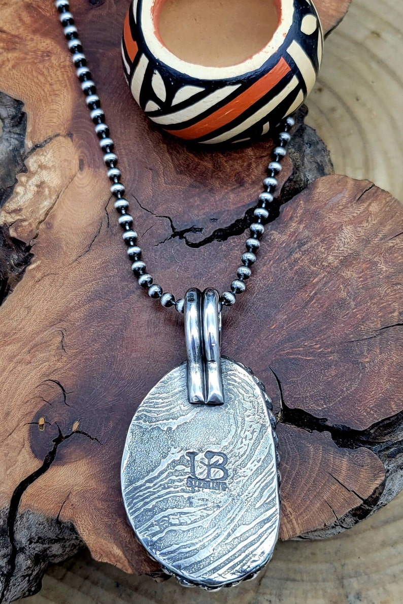 May include: A silver pendant necklace with a textured oval pendant. The pendant has a silver bail and the initials "UB" stamped on the back. The necklace is on a silver chain.