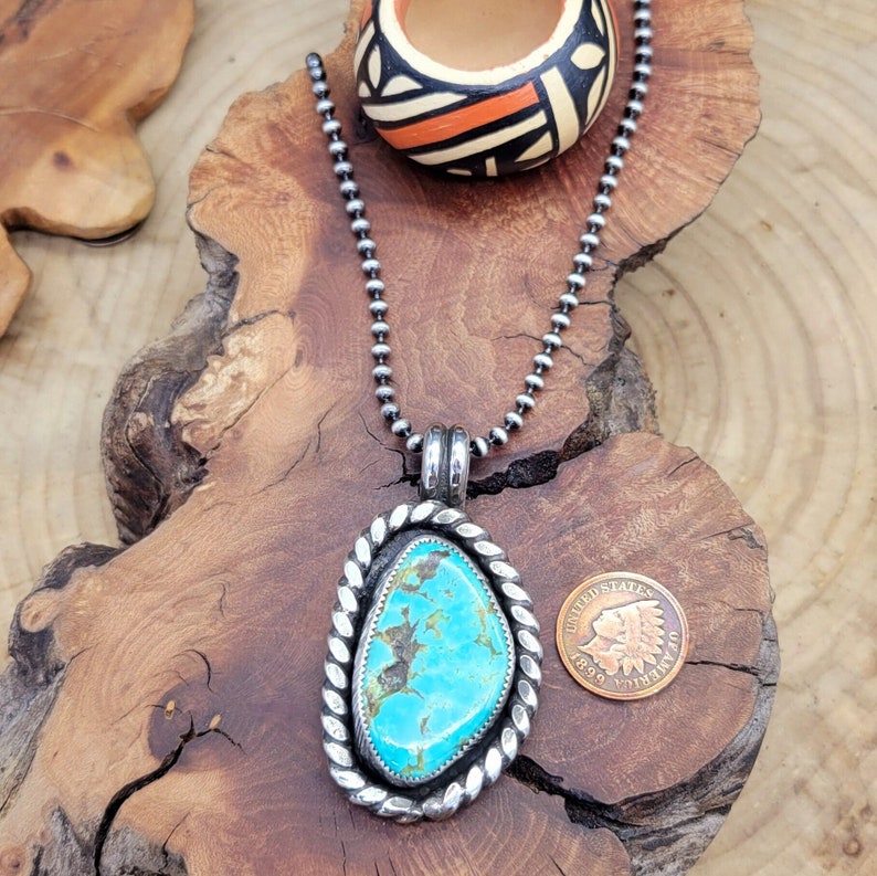 May include: A silver necklace with a turquoise stone pendant. The pendant is set in a silver frame with a rope design. The necklace is on a wooden surface with a small coin.