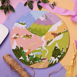 Puede incluir: Ilustración redonda y colorida que representa un paisaje montañoso con varios animales y elementos estacionales. El diseño presenta un zorro, un oso, un ciervo y un lobo, con picos nevados, árboles y una pequeña casa. La paleta de colores incluye rosa, verde, azul y amarillo.
