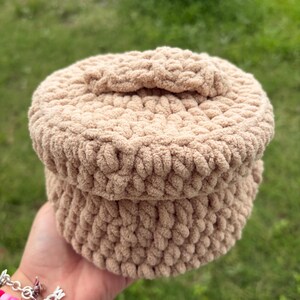 May include: A beige crocheted hat with a rounded crown and a small bow detail. The hat is made of a soft, textured yarn and appears to be handmade. The hat is held in a hand against a blurred green background.