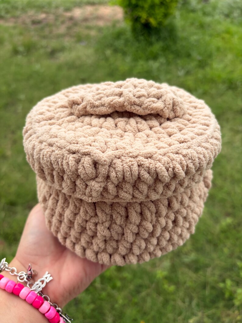 May include: A beige crocheted hat with a rounded crown and a small bow detail. The hat is made of a soft, textured yarn and appears to be handmade. The hat is held in a hand against a blurred green background.