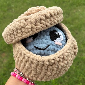 May include: A crocheted plush toy in a tan basket with a lid. The toy is blue with black and white eyes and a smiling mouth. The basket is tan and has a textured, woven appearance. A pink and silver bracelet is visible.