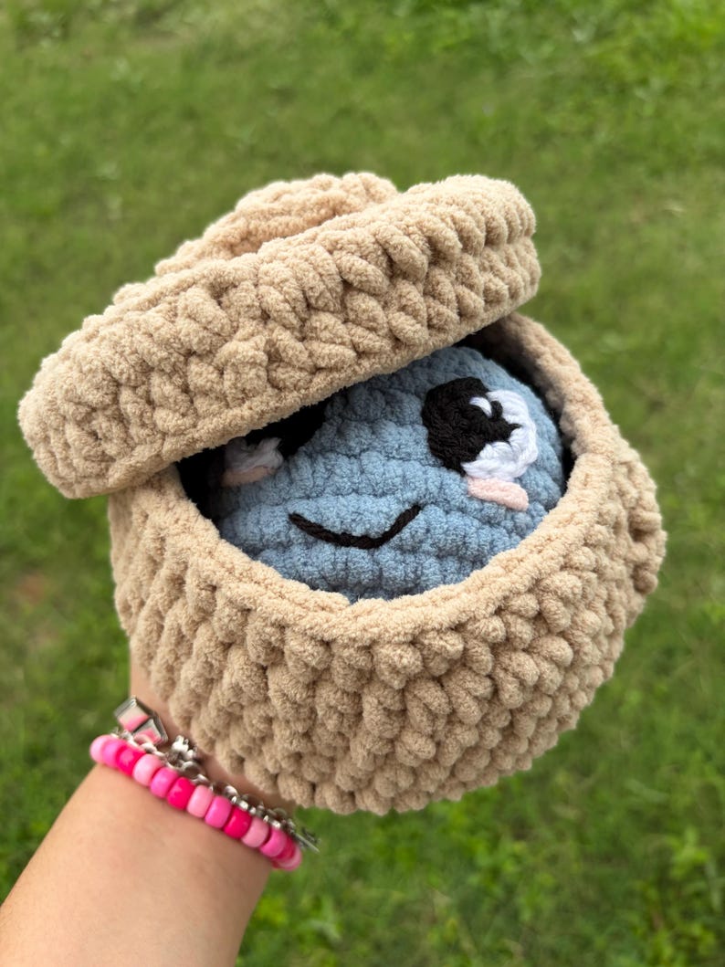 May include: A crocheted plush toy in a tan basket with a lid. The toy is blue with black and white eyes and a smiling mouth. The basket is tan and has a textured, woven appearance. A pink and silver bracelet is visible.