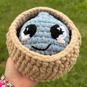 May include: A handmade, blue crocheted creature with black and white eyes and a smiling mouth, nestled in a tan crocheted basket. The plush toy is soft and cuddly, perfect for a child's room or as a unique gift.