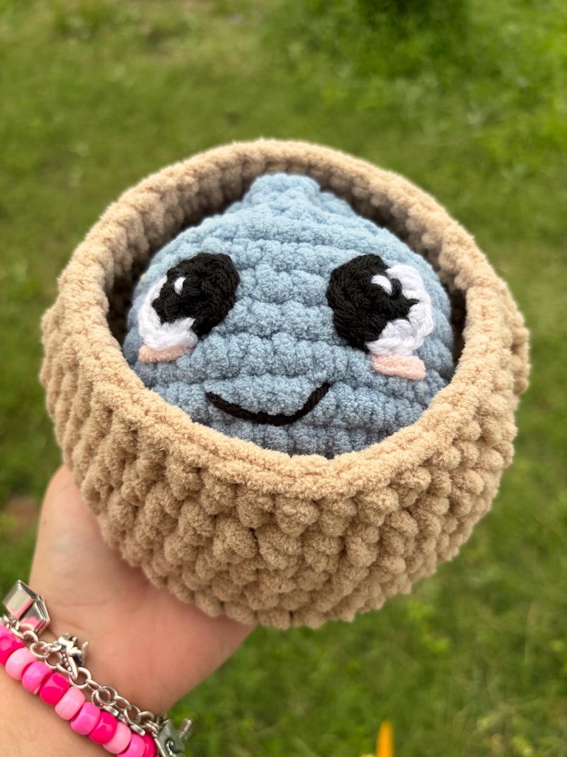 May include: A handmade, blue crocheted creature with black and white eyes and a smiling mouth, nestled in a tan crocheted basket. The plush toy is soft and cuddly, perfect for a child's room or as a unique gift.