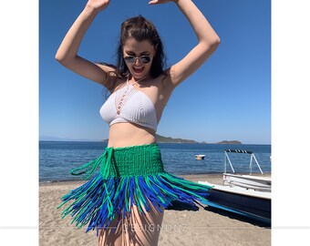 May include: A woman wearing a white crochet halter top and a green and blue fringed skirt poses on a beach. The skirt has a green waistband and blue and green fringe.
