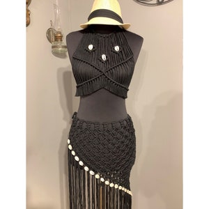 Pode incluir: Conjunto de top e saia de crochê preto com detalhes de conchas brancas. O top tem um design cruzado e a saia tem uma bainha com franjas.