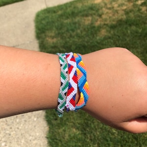Puede incluir: Tres pulseras de la amistad apiladas en una muñeca. Las pulseras están hechas de hilo de colores y presentan diferentes patrones. La pulsera superior es azul, naranja y amarilla. La pulsera del medio es roja, blanca y azul. La pulsera inferior es verde, blanca y gris.