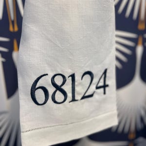 May include: A white linen hand towel with the embroidered number "68124" in navy blue. The towel has a stitched hem and is hanging in front of a navy blue patterned background.
