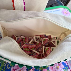 May include: A canvas tote bag with a zipper, filled with colorful mahjong tiles. The bag has a white exterior with red and purple scalloped trim. Pink dice and tiles are scattered on a blue floral patterned surface.