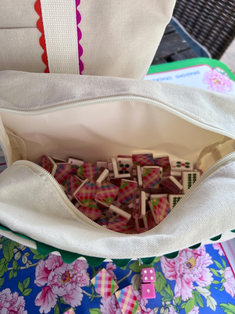 May include: A canvas tote bag with a zipper, filled with colorful mahjong tiles. The bag has a white exterior with red and purple scalloped trim. Pink dice and tiles are scattered on a blue floral patterned surface.